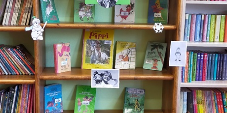 Powiększ grafikę: Regał biblioteczny, na którym wystawione są książki autorstwa Astrid Lindgren. W centralnym miejscu regału wisi karteczka z napisem „Była raz sobie…Astrid Lindgren” na dolnym brzegu kartki znajduje się okrągła grafika z czarno-białym wizerunkiem autorki. Po bokach regału są umieszczone wycinanki: bałwanek, śnieżynka, czarno-białe ksero zdjęcia Pipi Langstrumpf, Dzieci z Bullerbyn.