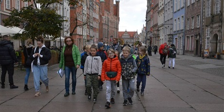 Powiększ grafikę: Barwna fotografia w kształcie prostokąta leżącego przedstawiająca grupę dziesięciorga dziewięcioletnich dzieci z dorosłą kobietą - przewodniczką miejską - na spacerze po ul. Długiej w Gdańsku. Grupa idzie środkiem ulicy, po obu stronach widać rzędy kamieniczek.