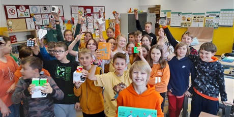 Powiększ grafikę: Wielobarwna grafika w kształcie prostokąta leżącego. W sali lekcyjnej, na środku stoją uczniowie biorący udział w konkursie układania kostki Rubika. Uczniowie trzymają w rękach kostki Rubika, a wyróżnieni uczniowie trzymają nagrody. 