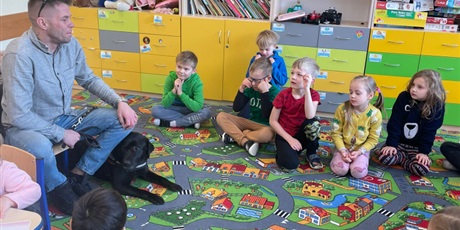 Powiększ grafikę: Barwna fotografia w kształcie prostokąta leżącego, przedstawiająca dzieci z oddziału przedszkolnego siedzące w kręgu na wykładzinie w sali lekcyjnej. Z dziećmi siedzi na krześle gość, obok którego leży pies przewodnik.