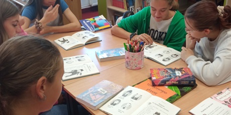 Powiększ grafikę: Wielobarwna fotografia w kształcie prostokąta stojącego. Na zdjęciu uczniowie podczas głośnego czytania w bibliotece szkolnej.