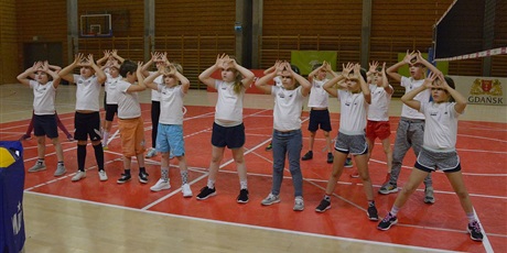 Powiększ grafikę: Barwna fotografia w kształcie prostokąta leżącego przedstawiająca 18-osobową grupę 9-letnich dzieci w strojach sportowych na sali gimnastycznej. Dzieci trzymają dłonie na wysokości czoła, pokazując prawidłowe ich ułożenie do odbicia siatkarskiego piłki. 
