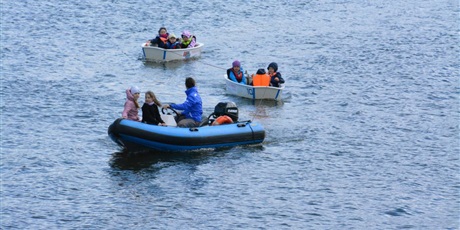 Powiększ grafikę: Barwna fotografia w kształcie prostokąta leżącego, przedstawiające zbiornik wodny, na środku którego płyną trzy łodzie powiązane ze sobą liną. Na łodziach siedzi ośmioro dziewięcioletnich dzieci i dwudziestokilkuletnia kobieta.