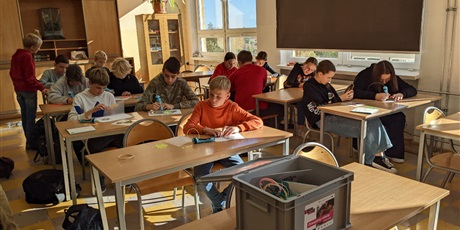 Powiększ grafikę: Barwna fotografia w kształcie prostokąta leżącego przedstawiająca uczniów z klasy 8F siedzących w ławkach, którzy wykorzystują długopisy 3D do tworzenia liści w kolorach jesieni. W tle sala lekcyjna.