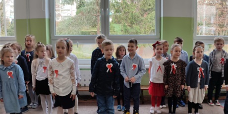 Powiększ grafikę: Wielobarwna grafika w kształcie prostokąta leżącego, przedstawiająca odświętnie ubranych uczniów klas 0 stojących na holu oddziałów przedszkolnych. Dzieci mają przypięte biało-czerwone kotyliony. 
