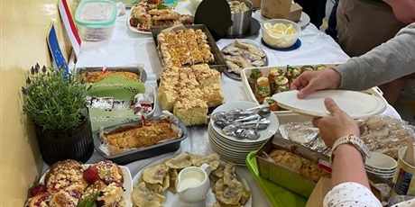 Powiększ grafikę: Barwna fotografia w kształcie prostokąta leżącego, przedstawiająca grupę osób stojących przy stołach z daniami kuchni polskiej. Na pierwszym planie pierogi, ciasto drożdżowe z owocami.