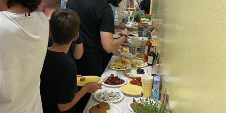 Powiększ grafikę: Barwna fotografia w kształcie prostokąta leżącego, przedstawiająca grupę osób stojących przy stołach z daniami kuchni hiszpańskiej, szwedzkiej, portugalskiej. Na pierwszym planie widać hiszpańskie wędzone kiełbasy, ser oraz inne tradycyjne przekąski.