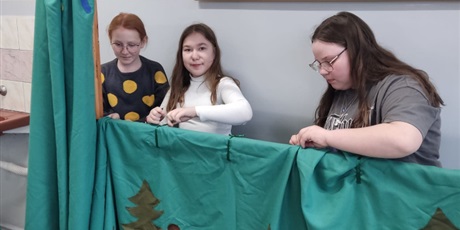 Powiększ grafikę: Prostokątne zdjęcie przedstawiające trzy uczennice klasy szóstej, przygotowujące elementy scenografii do przedstawienia teatralnego. Dziewczynki mocują zielony parawan z naszytymi choinkami. W tle ściana sali lekcyjnej.
