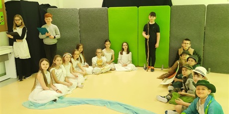 Powiększ grafikę: Prostokątne zdjęcie przedstawiające grupę teatralną. Osiem dziewczynek w białych sukniach siedzi po stronie lewej i sześciu chłopców w strojach barwy zielonej po stronie prawej. Między nimi stoi chłopiec ubrany na czarno. Trzyma w pionie długi, drewniany kij. W górnym, lewym rogu stoją dziewczynka z chłopcem. Ubrani są w stroje galowe. W dłoniach trzymają podkładki z klipem. Z przodu na podłodze błękitna narzuta. Z tyłu, w tle zielone materace.