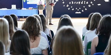 Powiększ grafikę: Barwna fotografia w kształcie prostokąta leżącego przedstawiająca uroczystość szkolną. Dyrektor stoi z mikrofonem i przemawia do grupy uczniów.