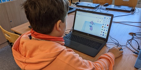 Powiększ grafikę: Barwna fotografia w kształcie prostokąta leżącego. Uczeń w pomarańczowej bluzie pracuje przy komputerze. Na ekranie monitora ucznia widać program Tinkercad. W tle szkolna pracownia komputerowa.