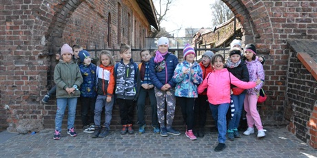 Powiększ grafikę: Barwna fotografia w kształcie prostokąta leżącego, przedstawiająca w centrum 13-osobową grupę dziewięcioletnich dzieci. Dzieci stoją na tle łukowatego otworu w murze starej budowli z czerwonej cegły.