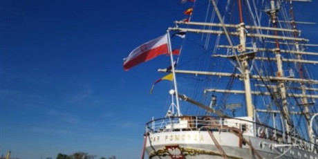 Powiększ grafikę: Barwna fotografia w kształcie prostokąta stojącego przedstawiająca żaglowiec "Dar Pomorza" zacumowany przy nabrzeżu w Gdyni. Na dziobie powiewa biało-czerwona flaga Polski.