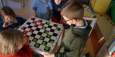 Powiększ grafikę: Barwna fotografia w kształcie prostokąta leżącego, przedstawiająca fragment szkolnego korytarza. Na środku znajduje się biały stolik z biało-czarną szachownicą przyklejoną na blacie. Na jej czarnych polach są ustawione plastikowe nakrętki w kolorze zielonym i czerwonym. Wokół stolika kuca lub klęczy pięcioro siedmioletnich dzieci. Grają w grę stolikową "warcaby".