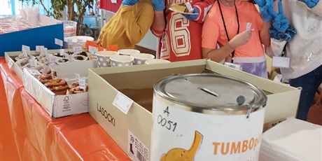 Powiększ grafikę: Wielobarwna grafika w kształcie prostokąta stojącego, przedstawiająca cztery wolontariuszki ubrane na pomarańczowo podczas obsługi kiermaszu. Z przodu skarbonka z napisem TUMBO pomaga oraz wypieki. 