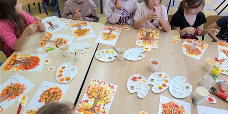 Powiększ grafikę: Wielobarwna fotografia w kształcie prostokąta leżącego przedstawiająca dzieci w świetlicy szkolnej, które siedzą wokół złączonych trzech ławek i malują farbami drzewa w kolorach jesieni.