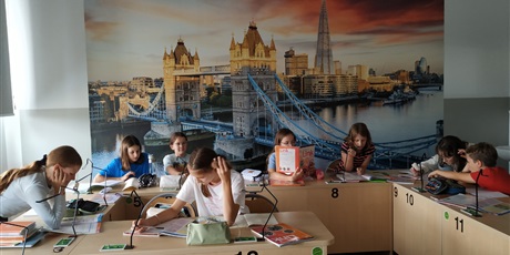 Powiększ grafikę: Wielobarwna fotografia w kształcie prostokąta leżącego. Na zdjęciu uczniowie klasy 5A, 5C i 5F podczas zajęć w pracowni języka angielskiego. Na ścianie fototapeta przedstawiająca Tower Bridge.